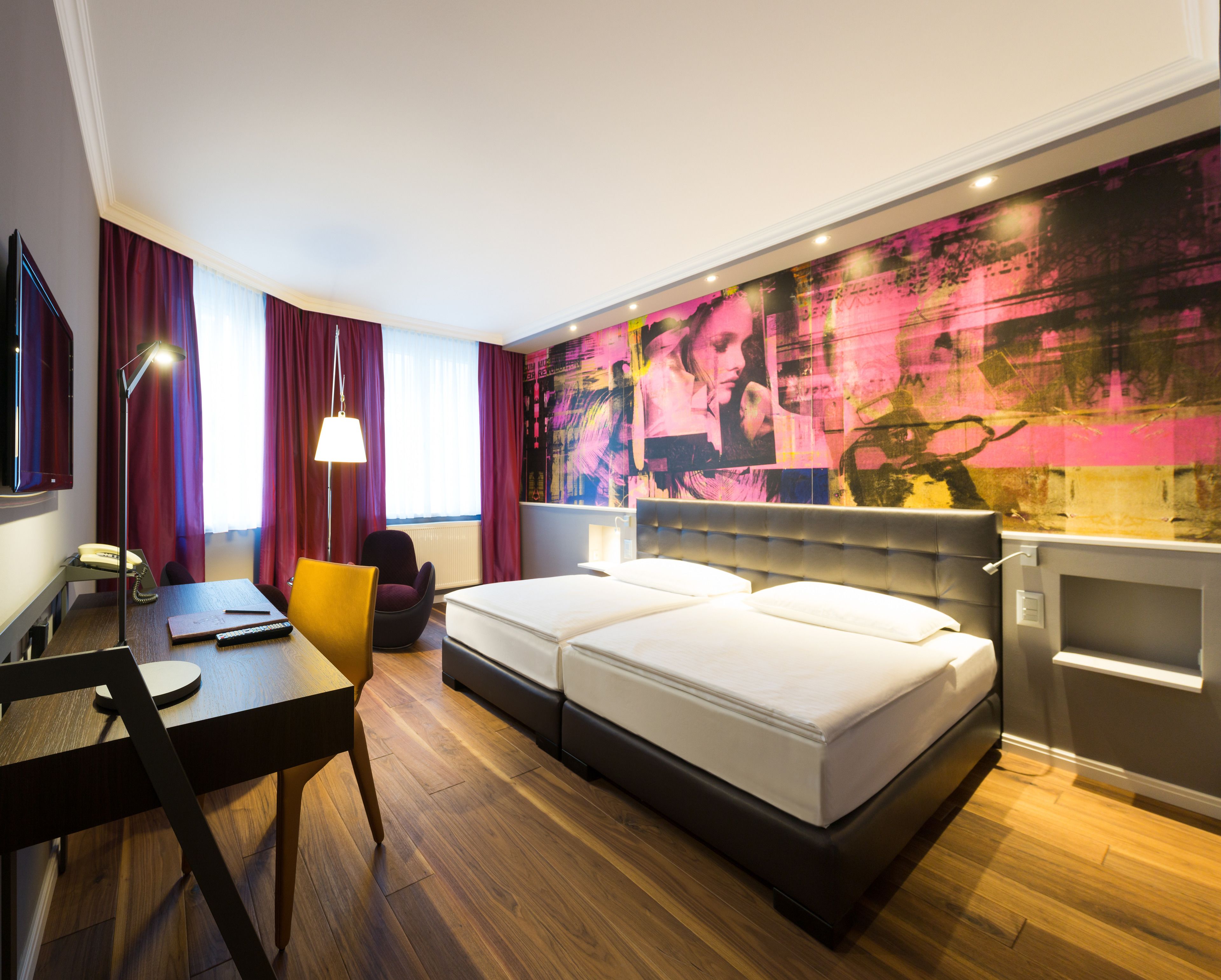 A hotel room with a bed, desk, chair, lamp, and a large abstract painting on the wall.