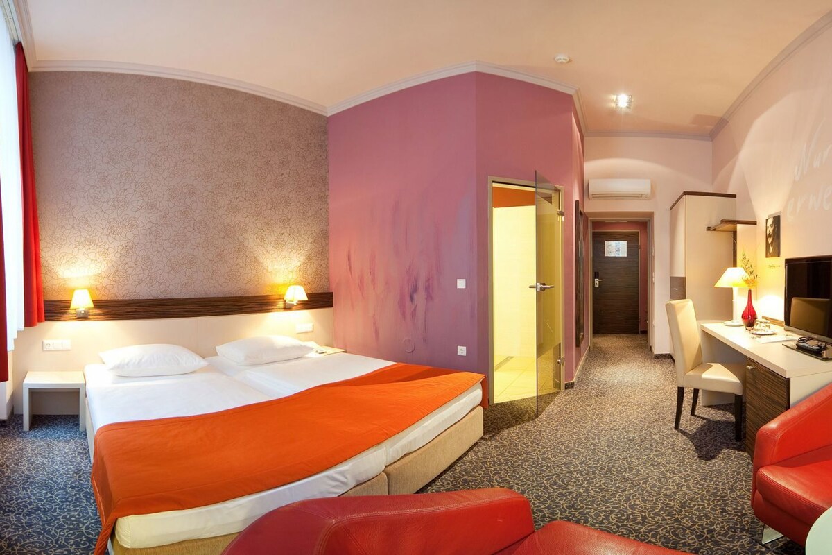 A hotel room with a bed, two bedside tables, a desk with a chair, a television, and a red sofa.