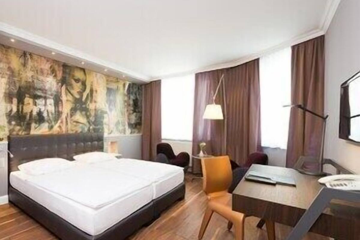 A hotel room with a large bed, a desk, a chair, a painting on the wall, and a window with curtains.
