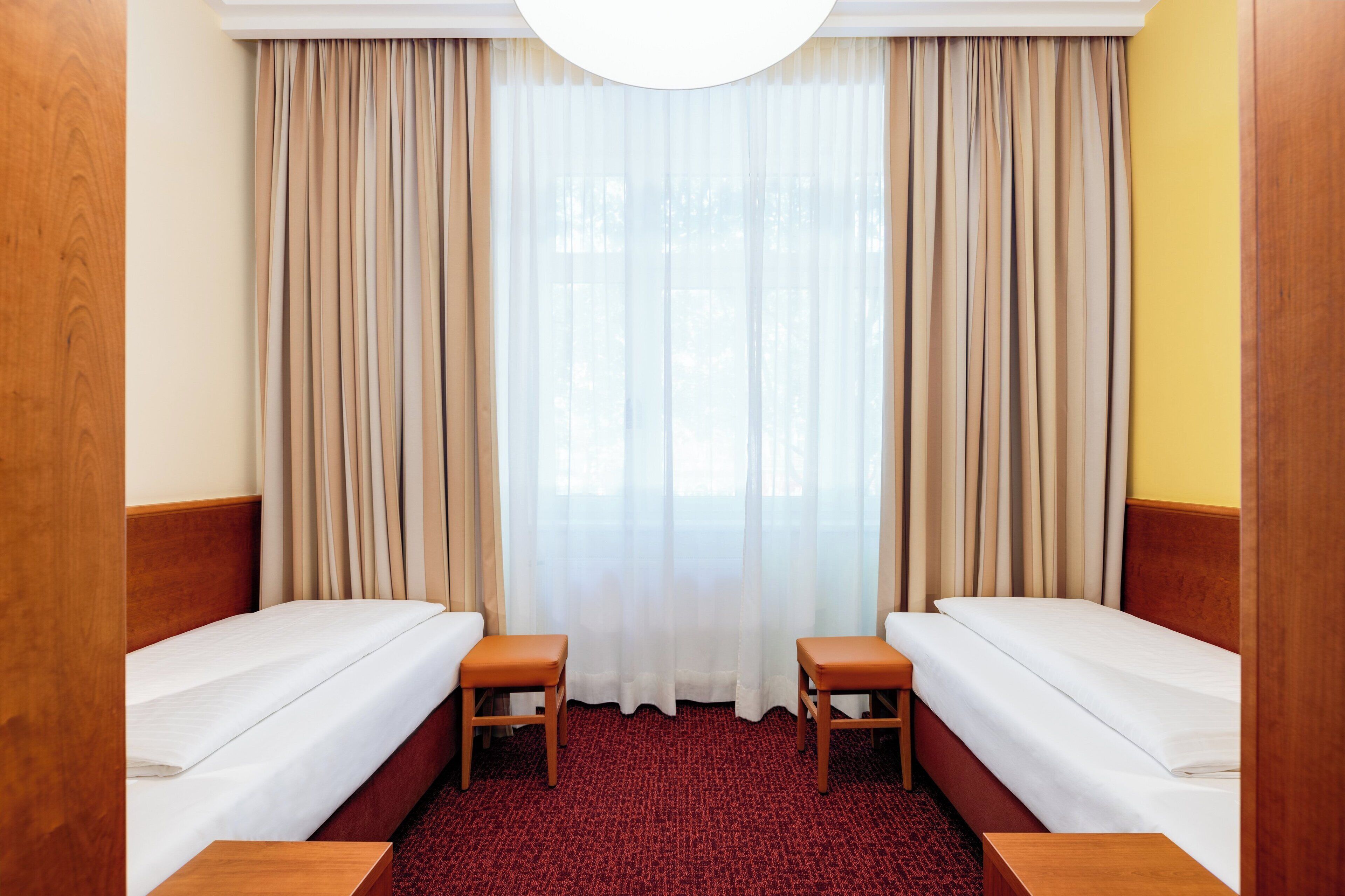 Ett hotellrum med två separata sängar, en sänggavel i trä, en röd matta och ett stort fönster med tunna, genomskinliga gardiner.