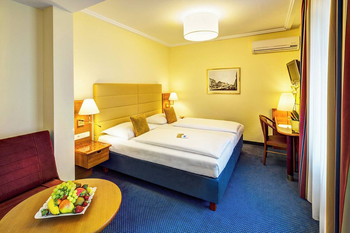 A hotel room with two beds, a dining table with fruit, a red sofa, and a desk with a lamp.