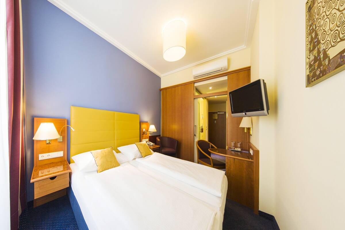 A hotel room with a bed, bedside tables, a chair, a desk, a television, and an air conditioning unit.