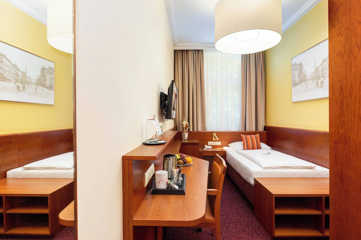 A hotel room with two single beds, a wooden desk with a coffee maker, a TV, and a large window with curtains.