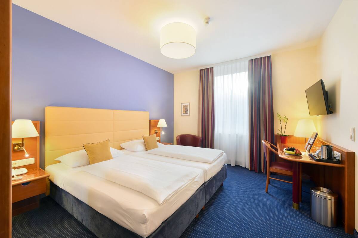 A hotel room with two beds, a desk with a chair, a TV, and a small trash bin.