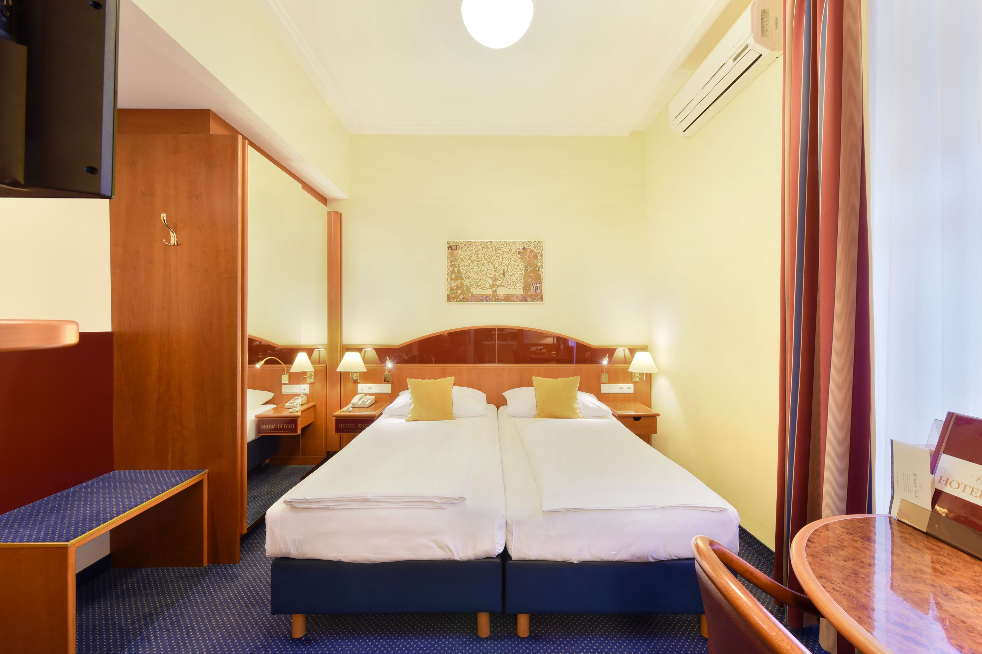 Una camera d'albergo con due letti, una scrivania, una sedia, un comodino, una lampada e un quadro appeso al muro.