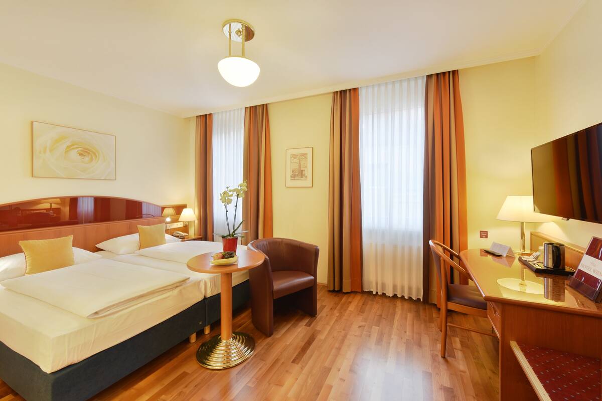 A hotel room with a bed, two bedside tables, a desk, a chair, a small table with a plant, and a large window with curtains.