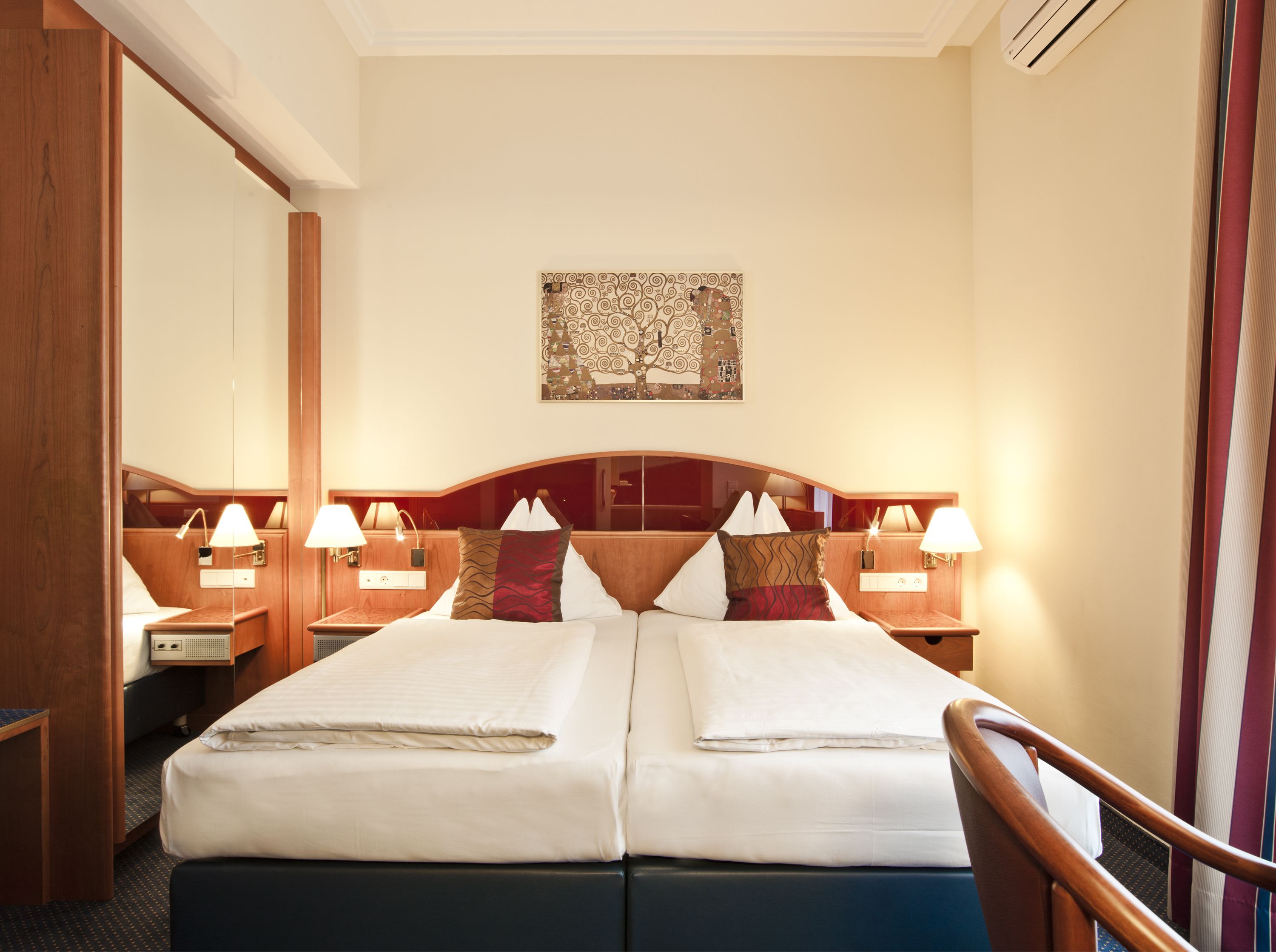 Quarto de hotel com uma cama de casal, duas mesas de cabeceira com luminárias, uma cabeceira com painel decorativo e um espelho.