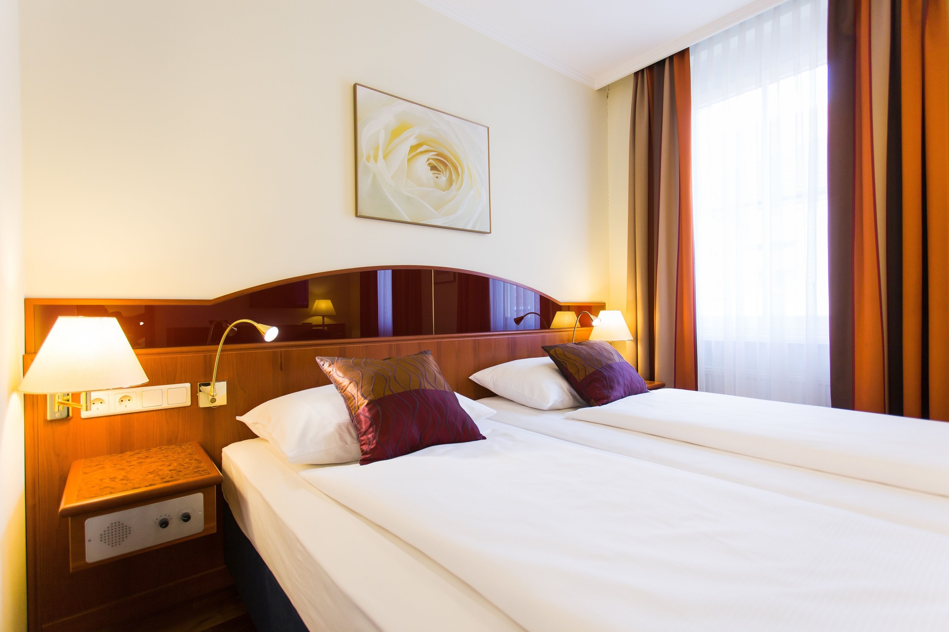 Habitación de hotel con dos camas, una mesita de noche con lámpara, una obra de arte en la pared y una ventana con cortinas.