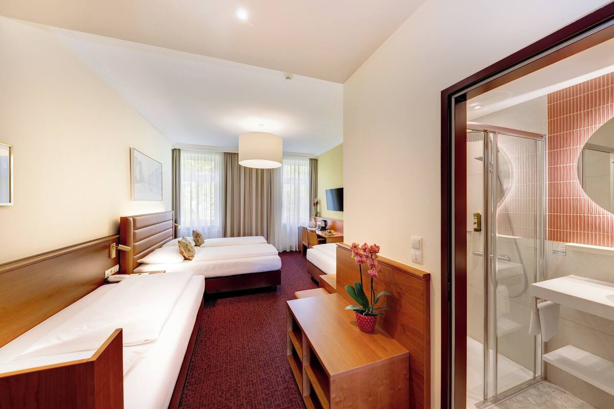 A hotel room with two beds, a desk, a chair, a potted orchid, and a glass-enclosed shower area.