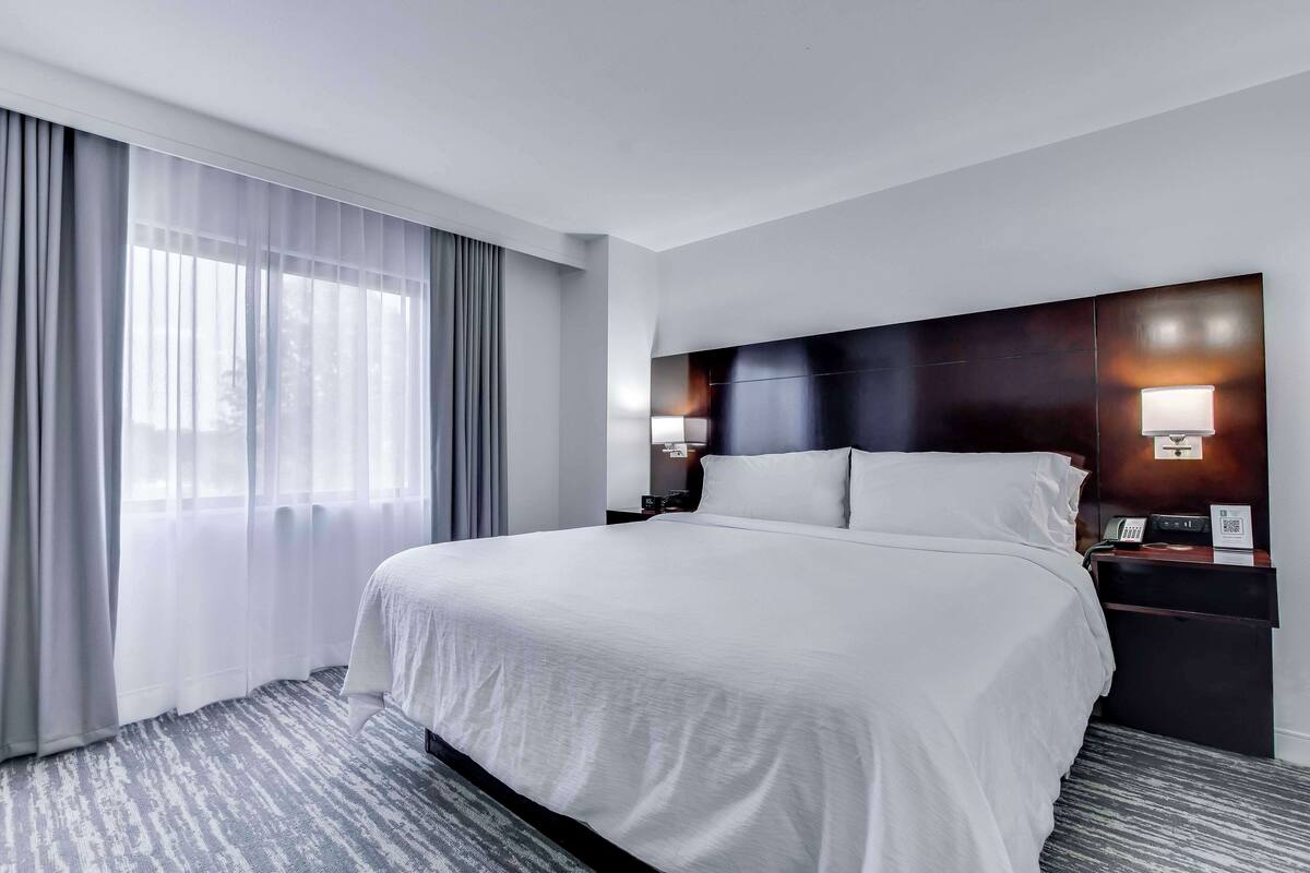 Une chambre d’hôtel avec un grand lit, une table de chevet, une lampe et une vue sur l’extérieur à travers des rideaux légers.