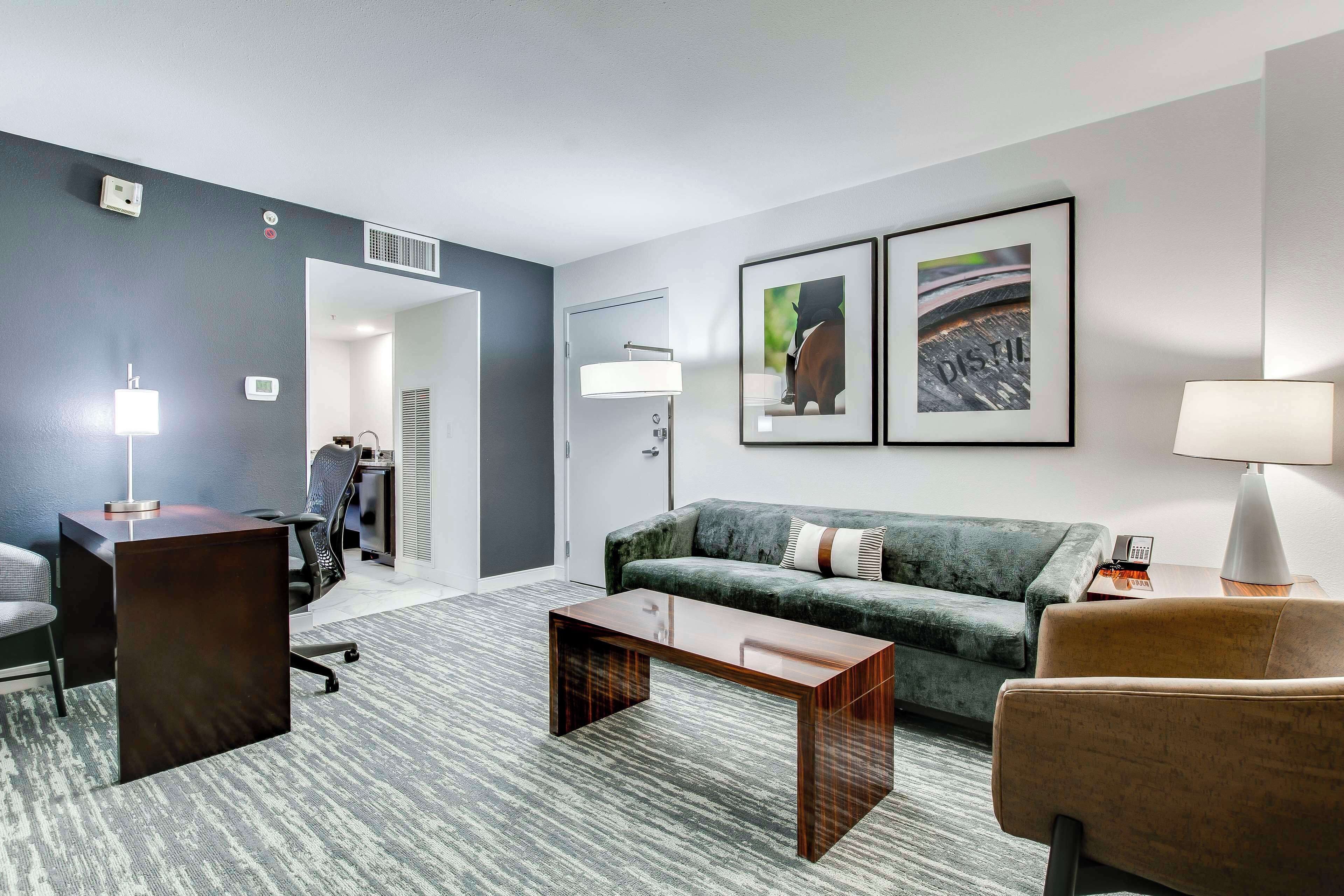 A modern hotel room with a dark grey wall, a grey carpet, a wooden coffee table, a green sofa, a brown armchair, and two framed pictures on the wall.