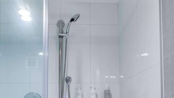 A modern bathroom with a glass shower enclosure, a handheld showerhead, a handheld shower attachment, and a small table with a towel.