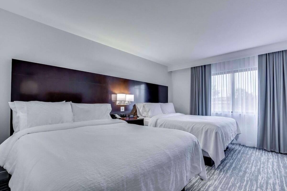 Une chambre d’hôtel avec deux lits, une table de chevet, une lampe et une grande fenêtre avec des rideaux.