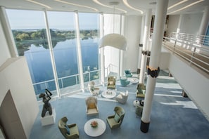 Un amplio salón con vistas panorámicas a un lago y al paisaje urbano, con mobiliario moderno y una gran lámpara colgante blanca.