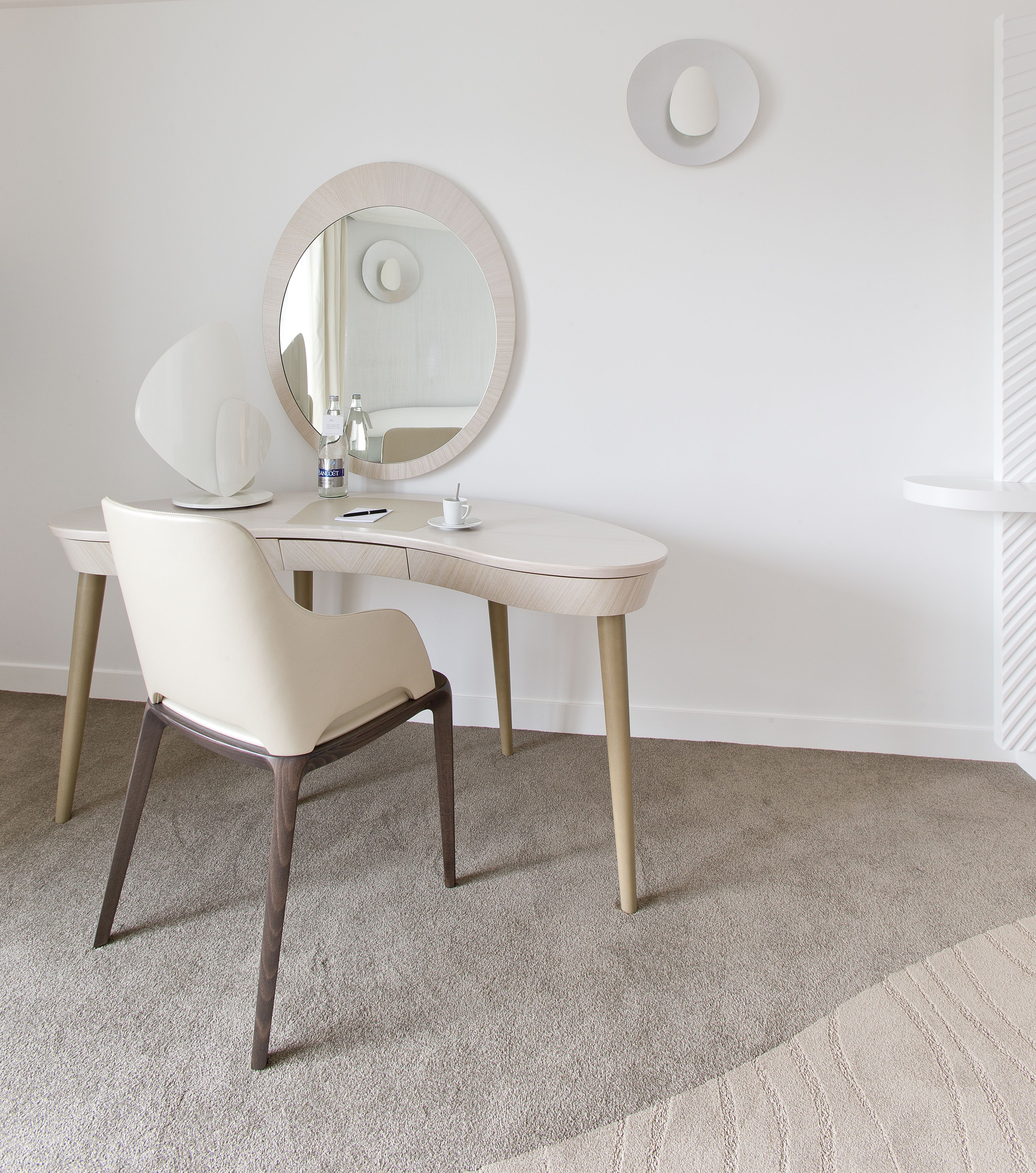 A minimalist room with a white vanity, a chair, a round mirror, and a small table with a cup and a bottle.