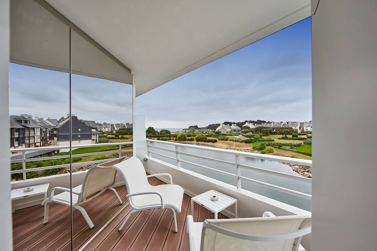A balcony with white chairs and a small table, overlooking a residential area with houses and greenery.