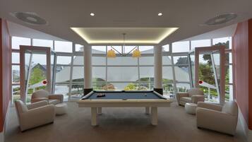 A pool table in a room with large windows, white walls, and beige furniture.