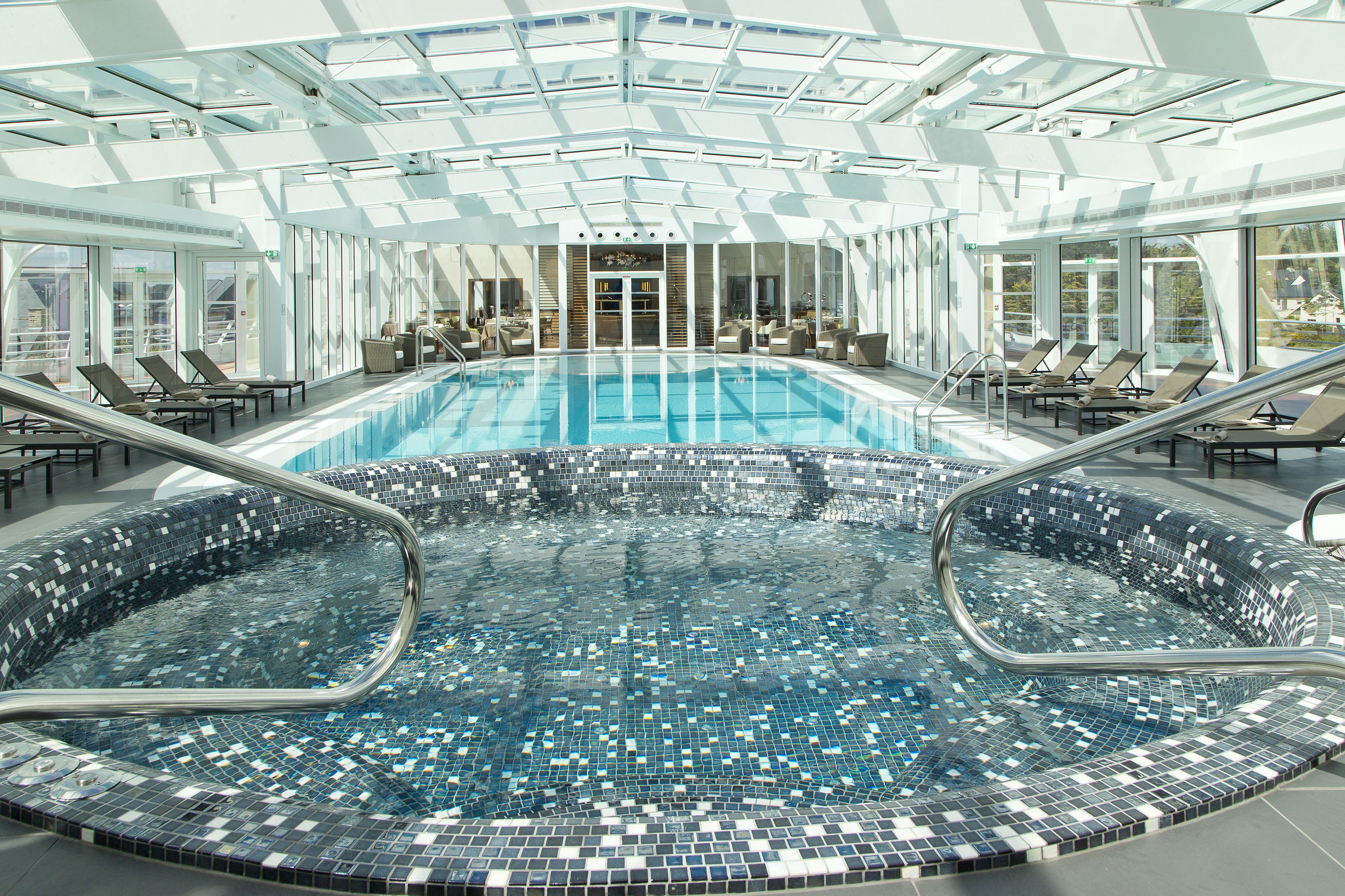 Une grande piscine intérieure aux bords carrelés, entourée de chaises longues et dotée d’un plafond en verre.