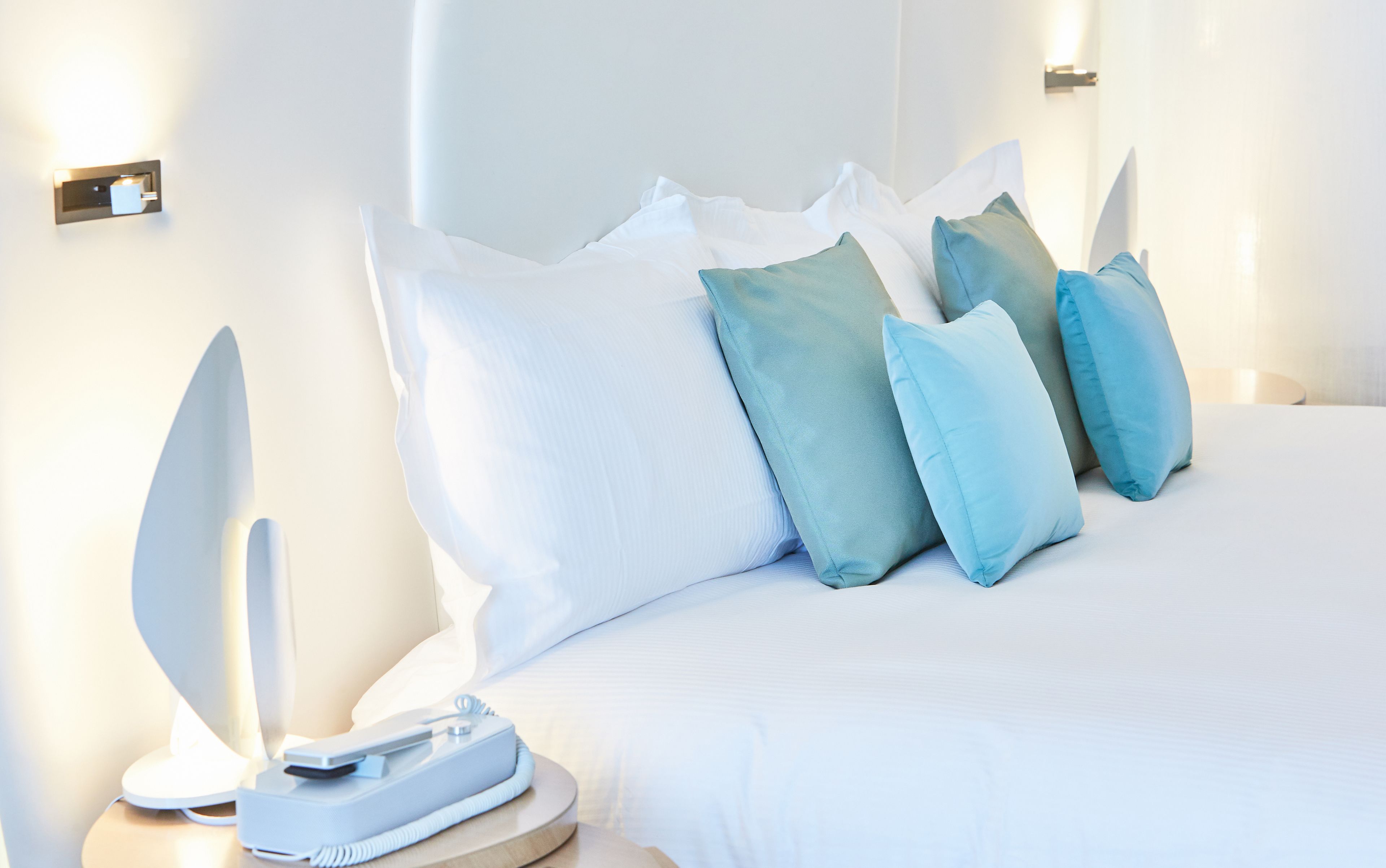 A neatly made bed with white and teal pillows, a bedside table with a lamp, and a wall-mounted light fixture.
