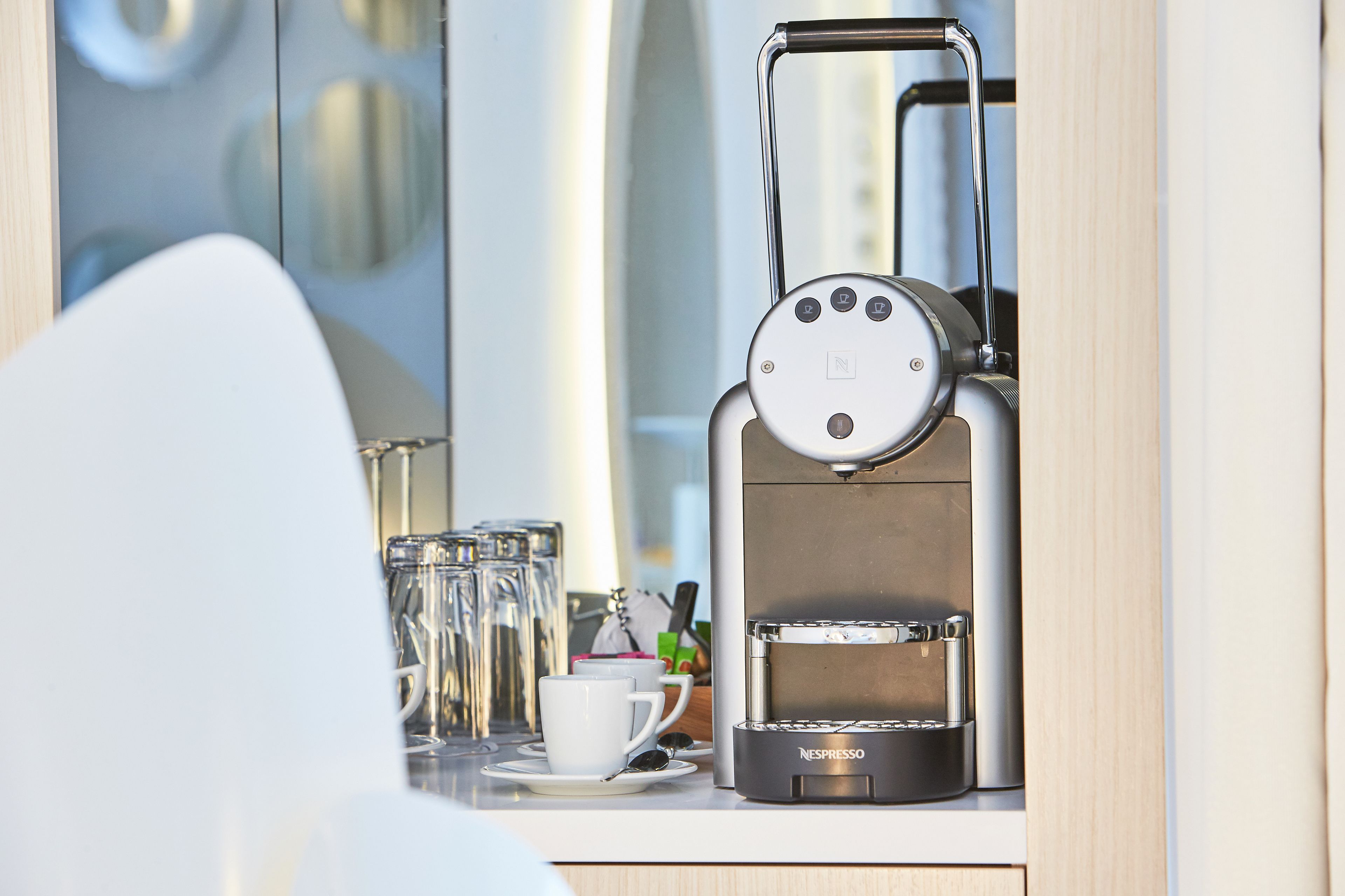 Nespresso-kahvinkeitin tasolla, sen vieressä kuppi ja aluslautanen, laseja ja pussi.
