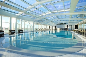 An indoor swimming pool with a glass ceiling, sun loungers, and a view of the city.