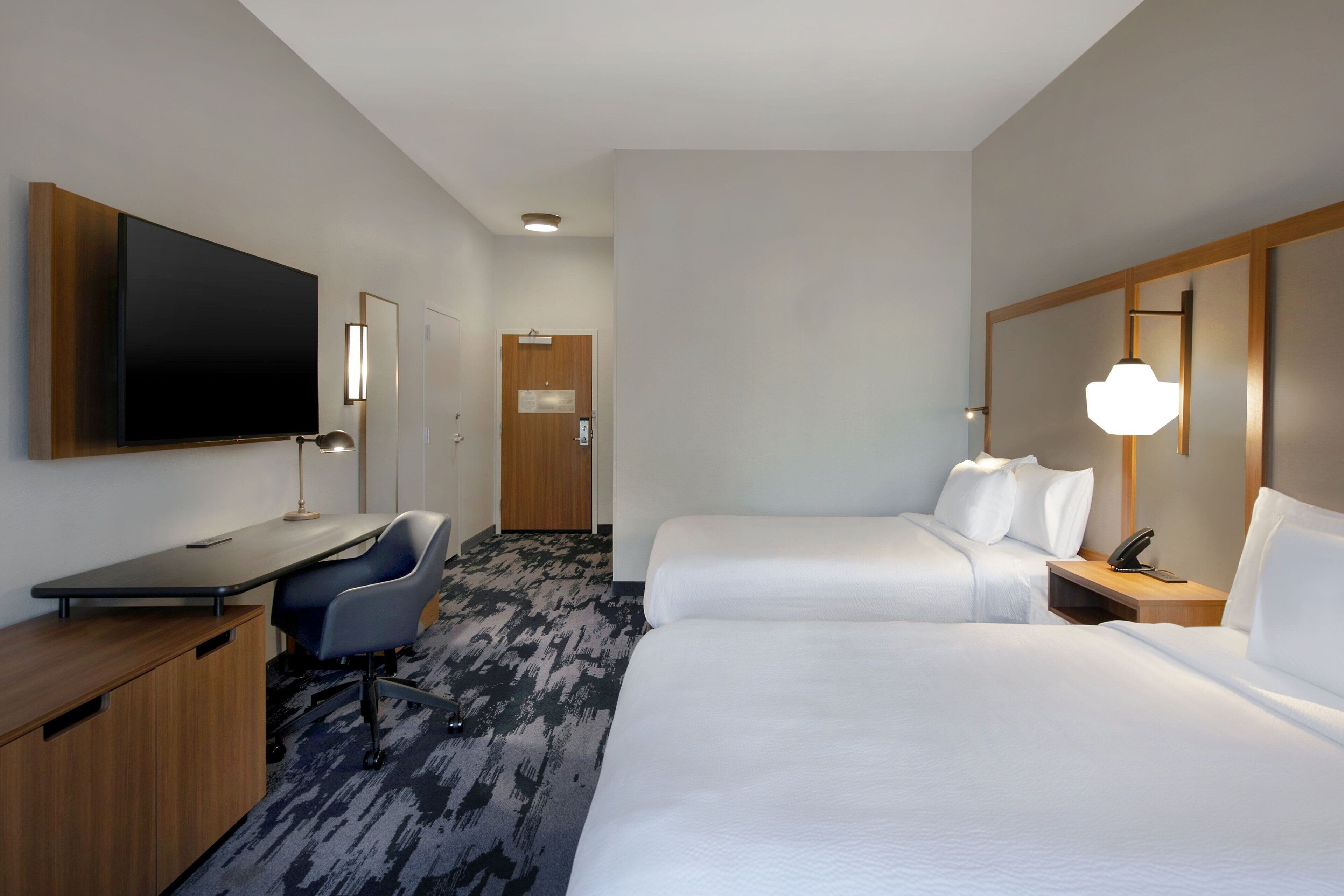 Habitación de hotel con televisión de pantalla plana, escritorio, dos camas, una silla, una lámpara y una puerta.
