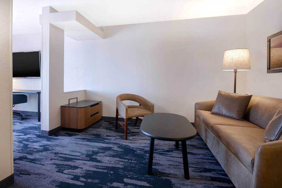 Una habitación de hotel moderna con sofá, una mesita, una silla, un escritorio y una lámpara, además de un televisor de pantalla plana.