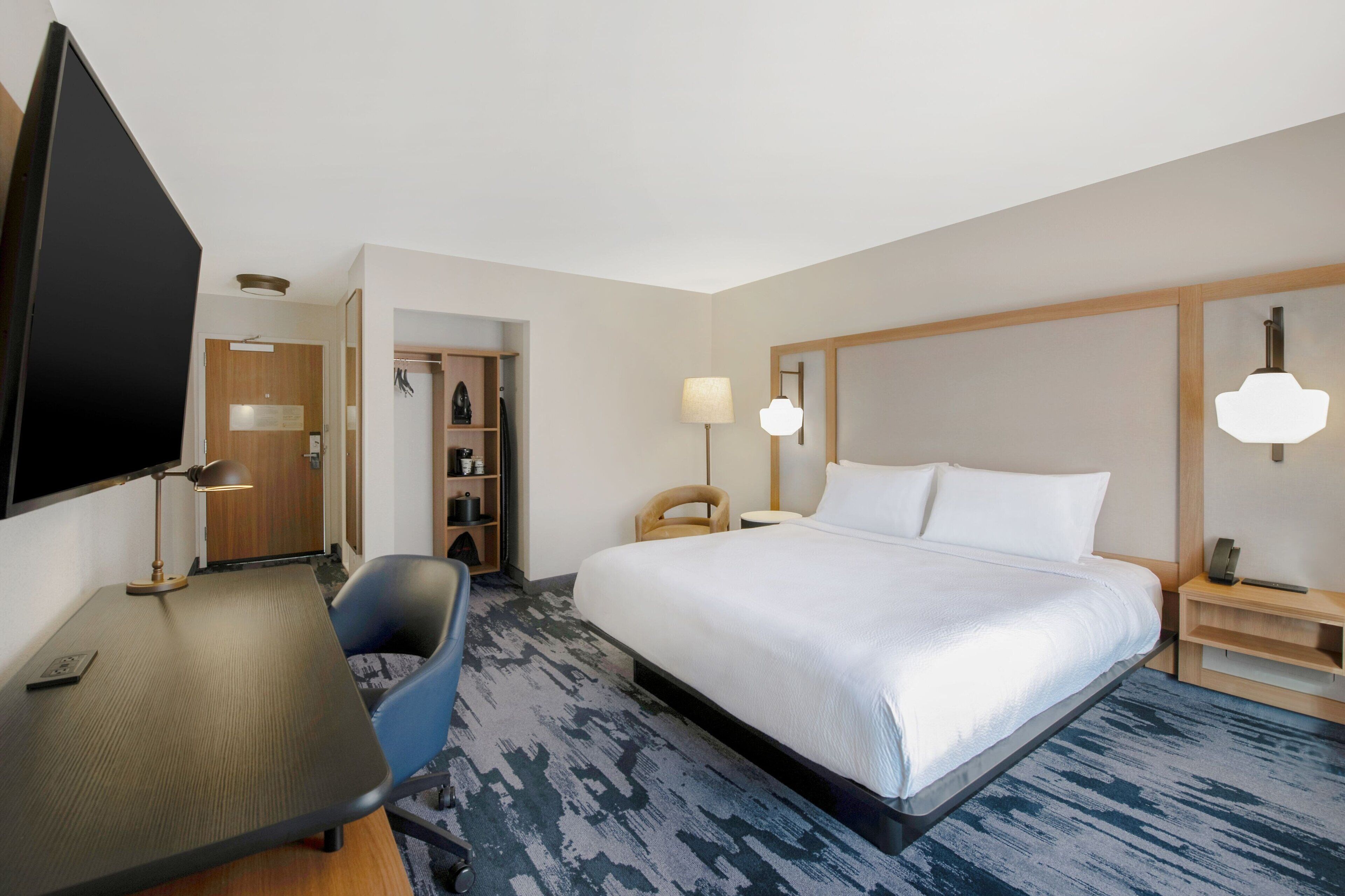 A hotel room with a bed, a desk with a chair, a TV, a lamp, and a nightstand.