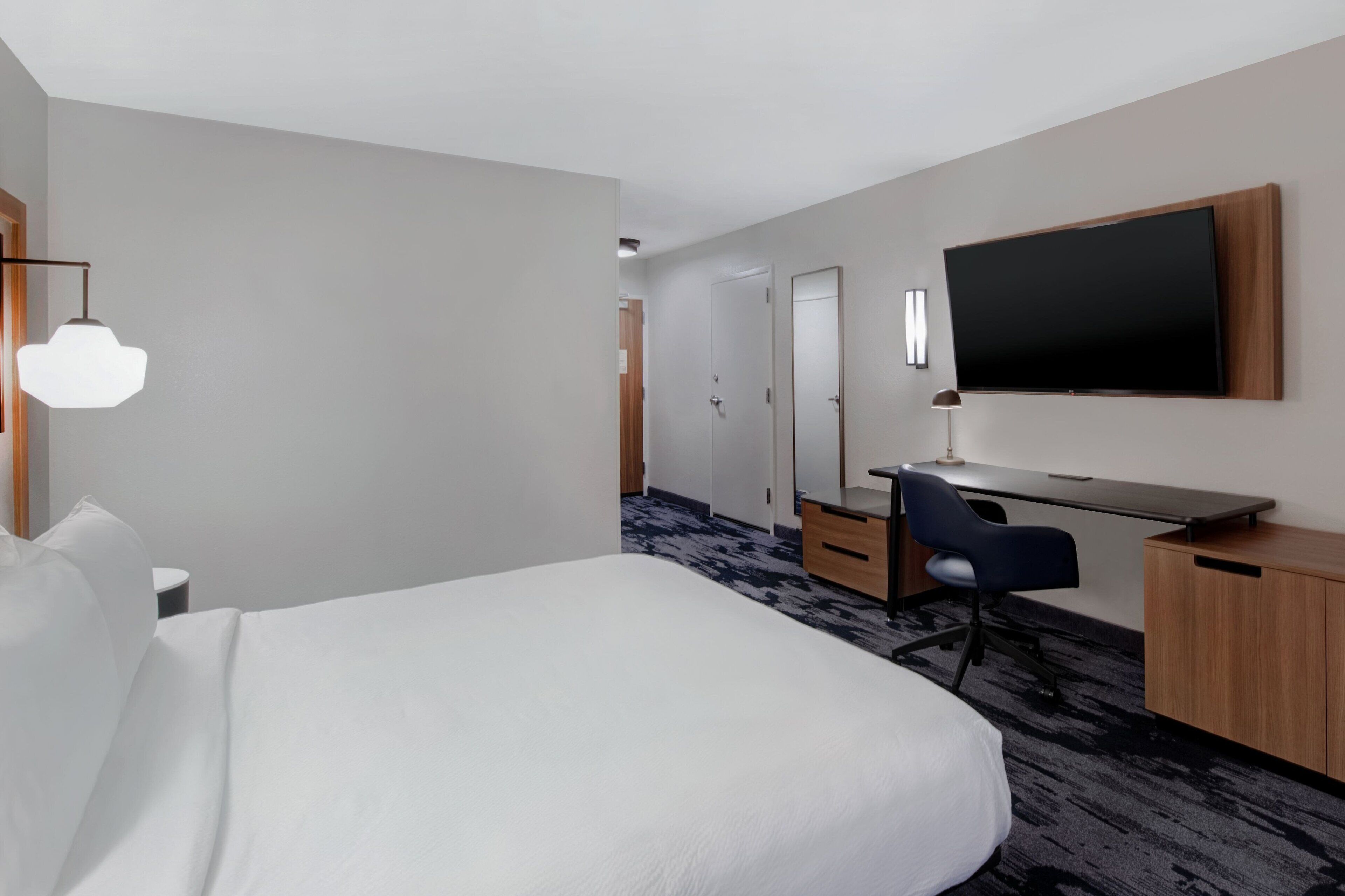 A hotel room with a large bed, a desk with a chair, a flat-screen TV mounted on the wall, and a small table with a lamp.
