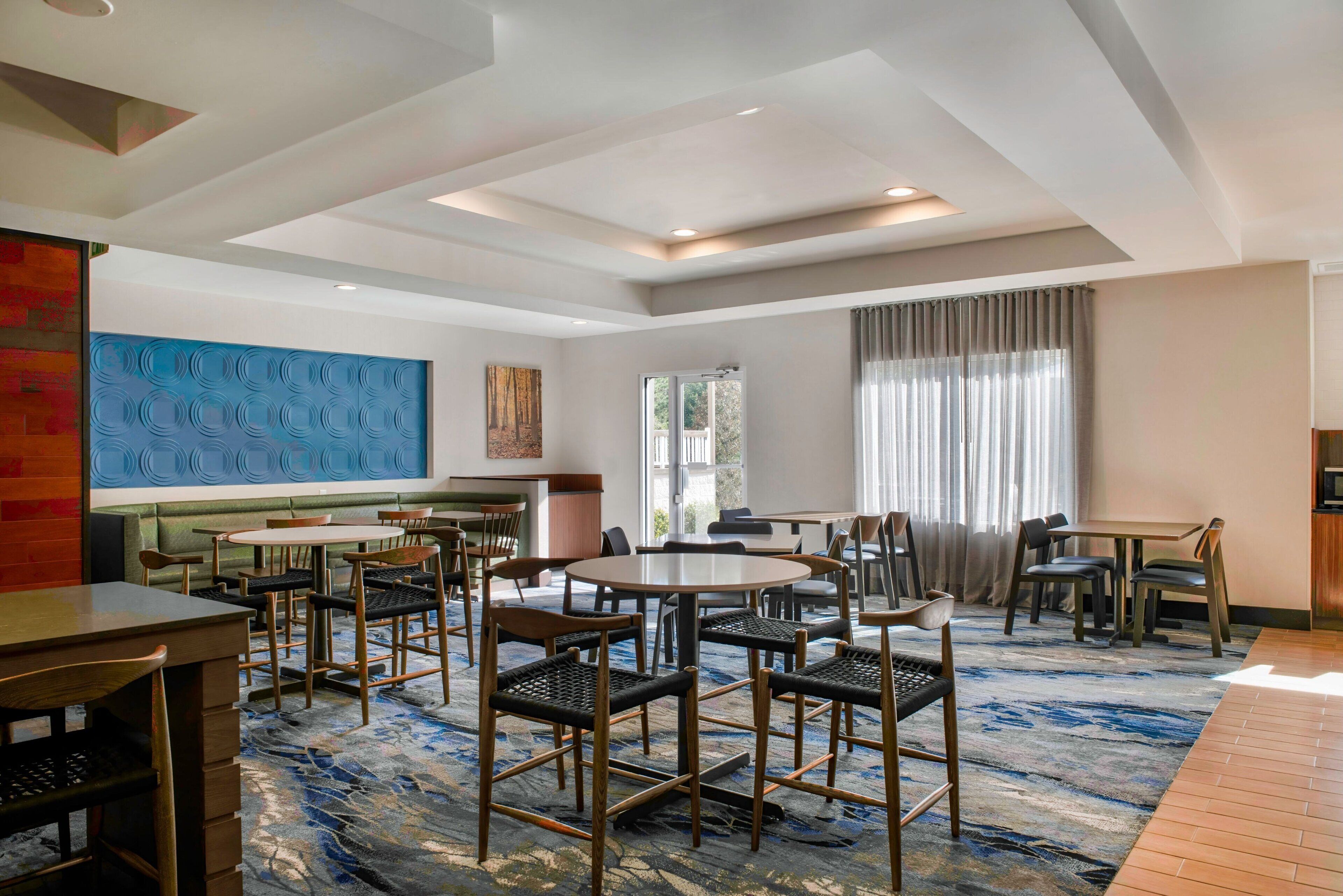 A modern hotel lobby with round tables, black and wooden chairs, a blue abstract wall art, and a large window with curtains.