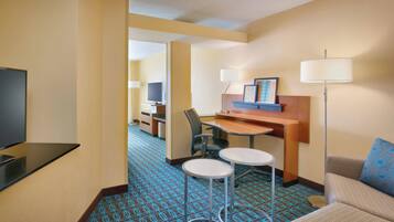 A hotel room with a TV, a desk with a chair, two small tables, a sofa with a patterned pillow, and a patterned carpet.