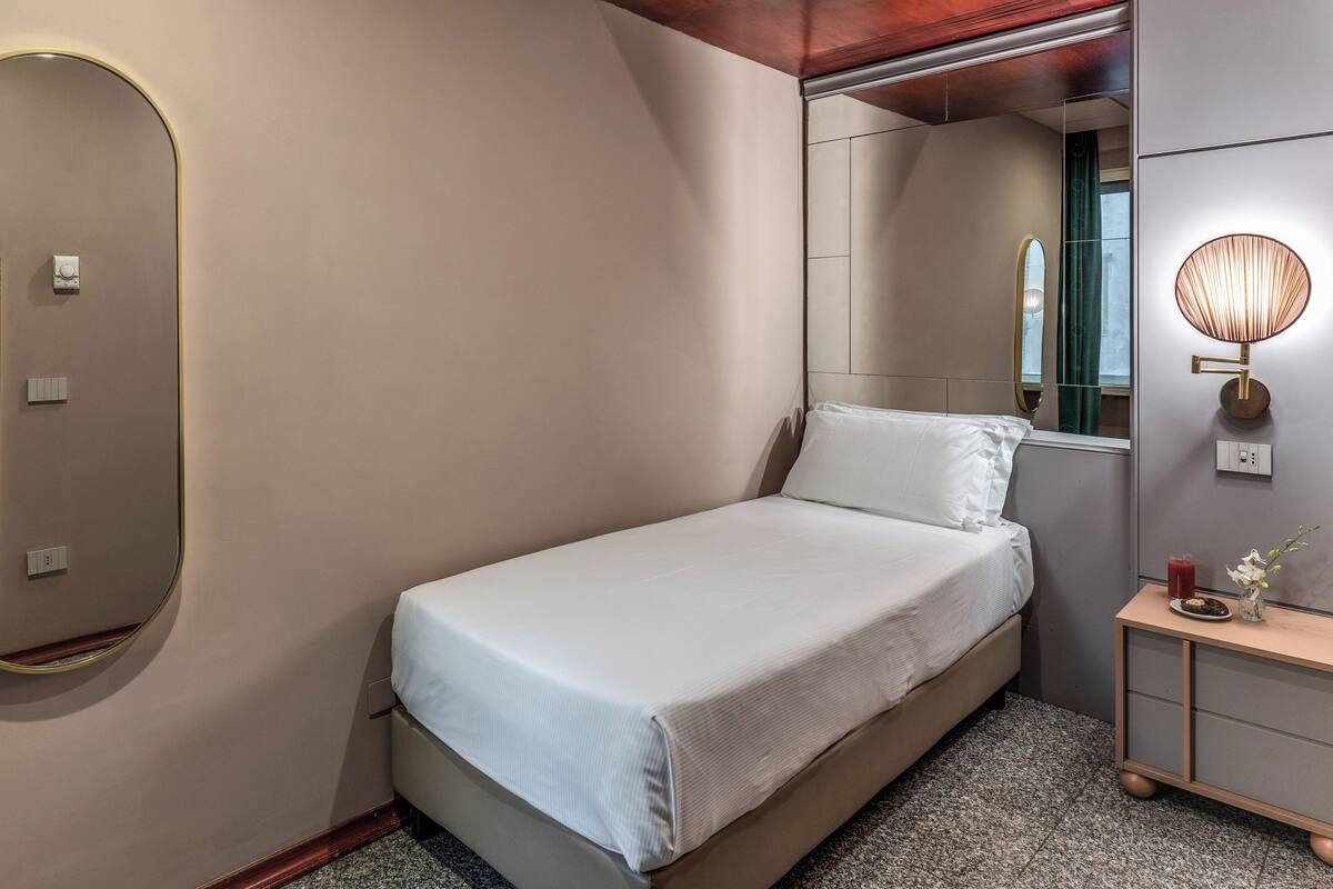A neatly made bed in a hotel room with a large mirror, a bedside table with a lamp, and a small vase with flowers.