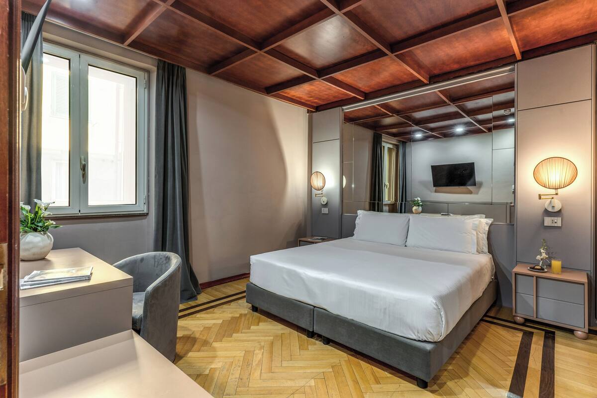 A modern bedroom with a large bed, a desk, and a chair. There is a TV mounted on the wall and a window with curtains.