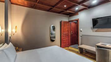 Habitación de hotel con cama, mesita de noche, espejo, puerta de madera, banco y televisor fijado a la pared.
