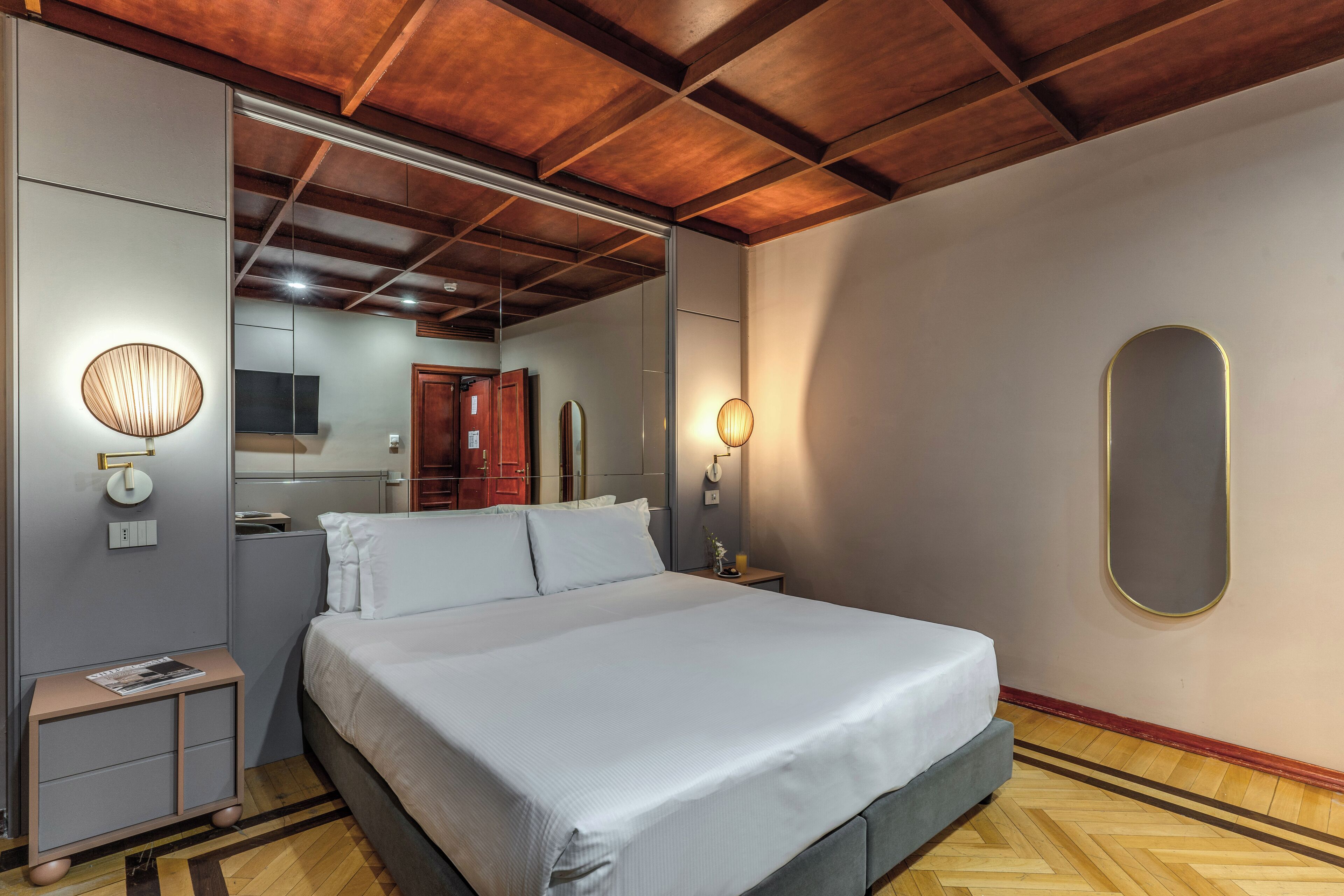 Un dormitorio moderno con una cama grande, vigas de madera en el techo, un espejo y una mesita de noche con lámpara.