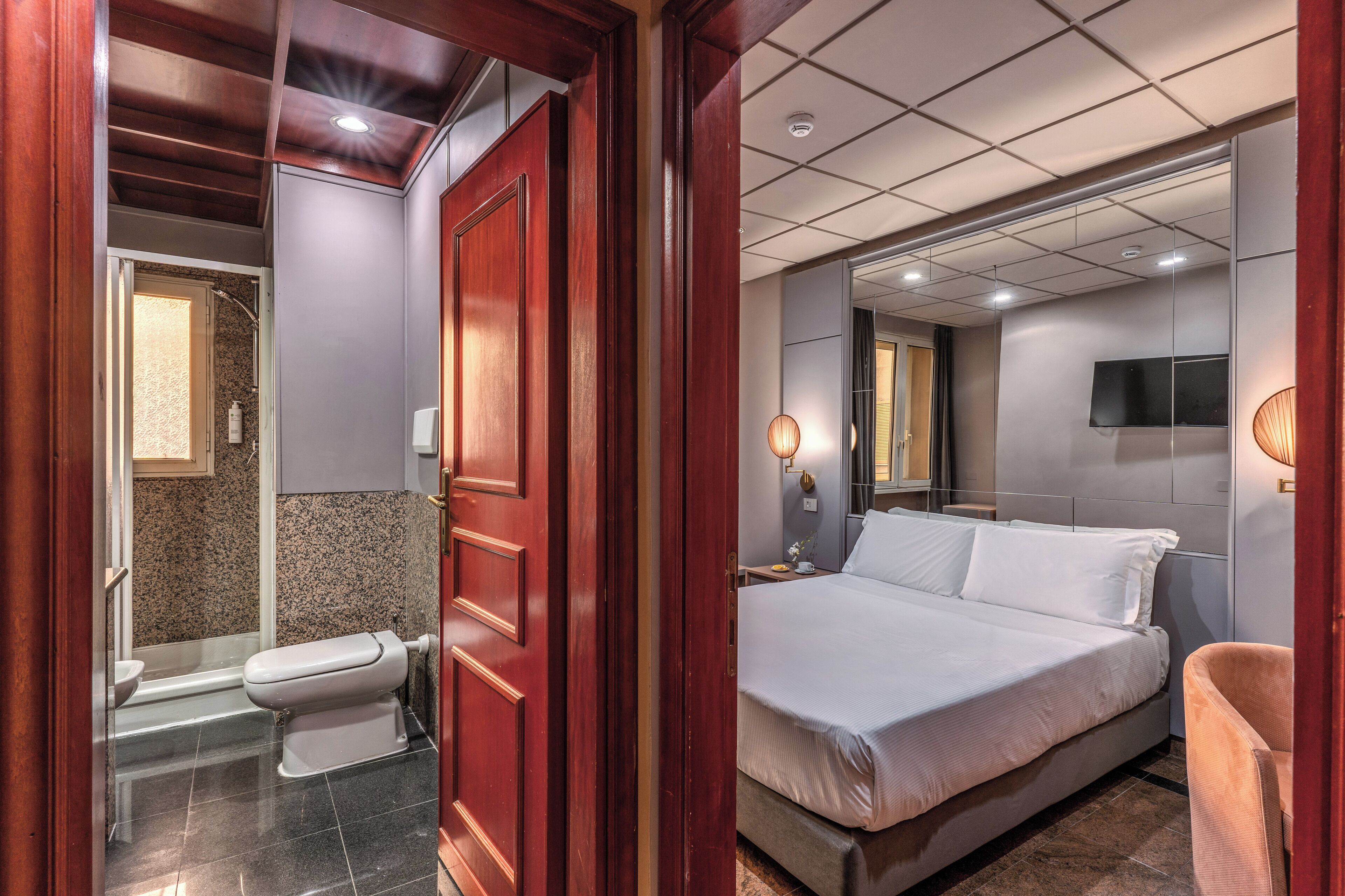 A hotel room with a wooden door, a shower, a toilet, a bed, a TV, and a nightstand with a lamp.