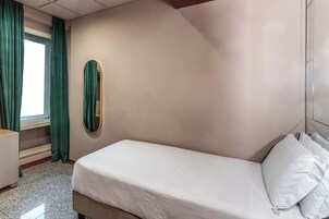 Un letto singolo con biancheria da letto bianca in una stanza con un'ampia finestra, tende verdi e uno specchio a parete.