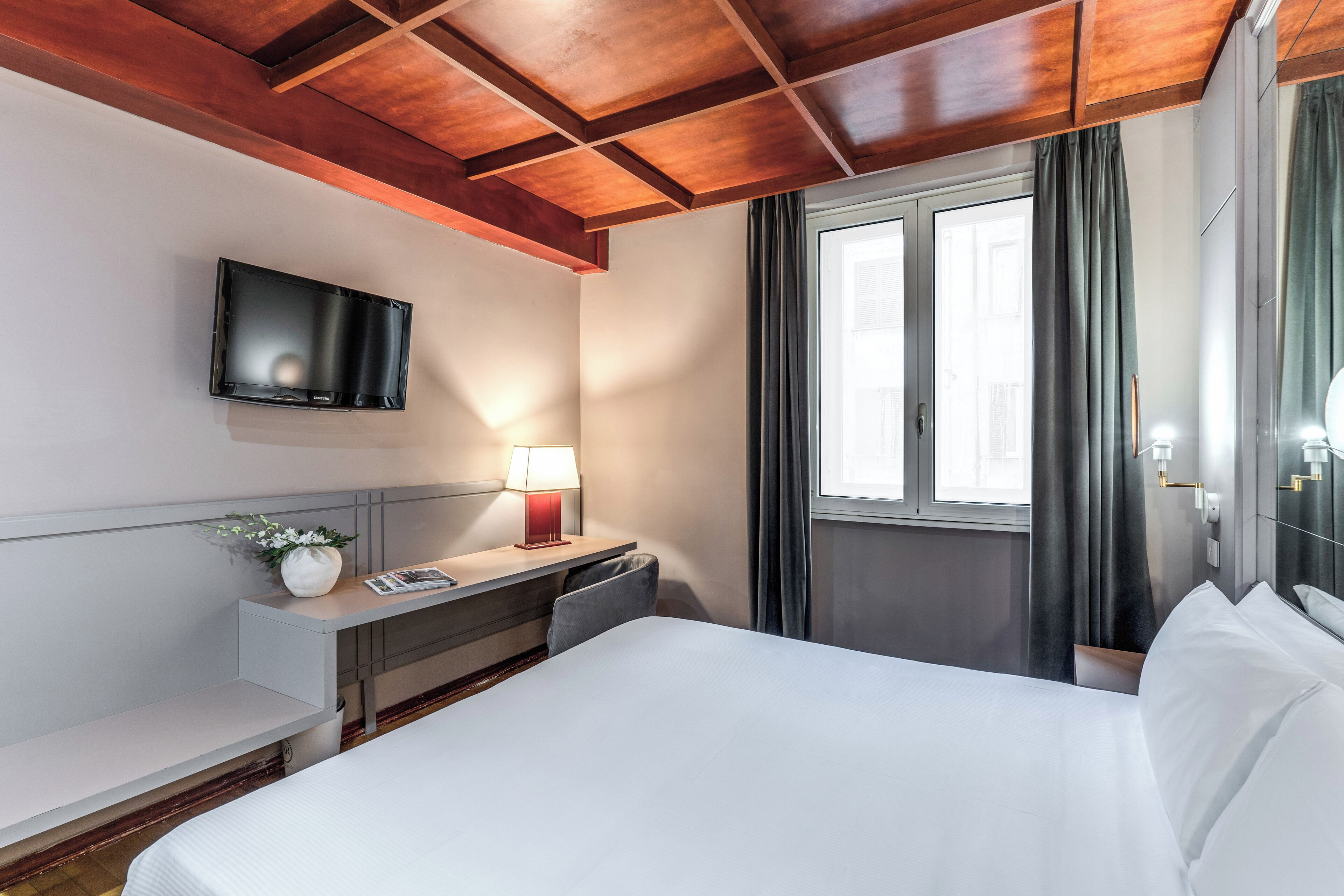 Habitación de hotel con cama, escritorio con lámpara, televisor de pantalla plana montado en la pared y ventana con cortinas.