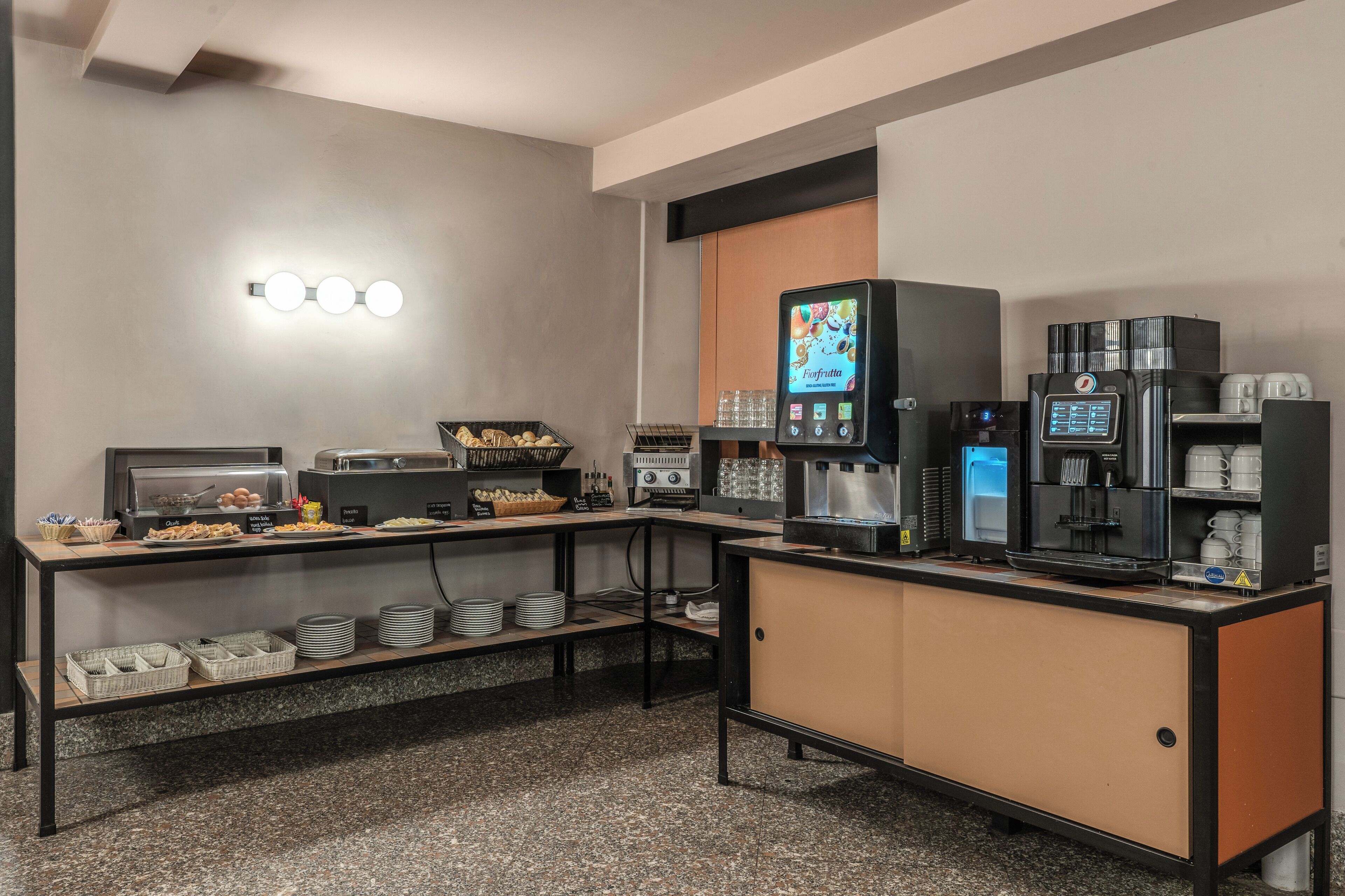 A self-service breakfast area with a coffee machine, tea station, and various food items on a counter.