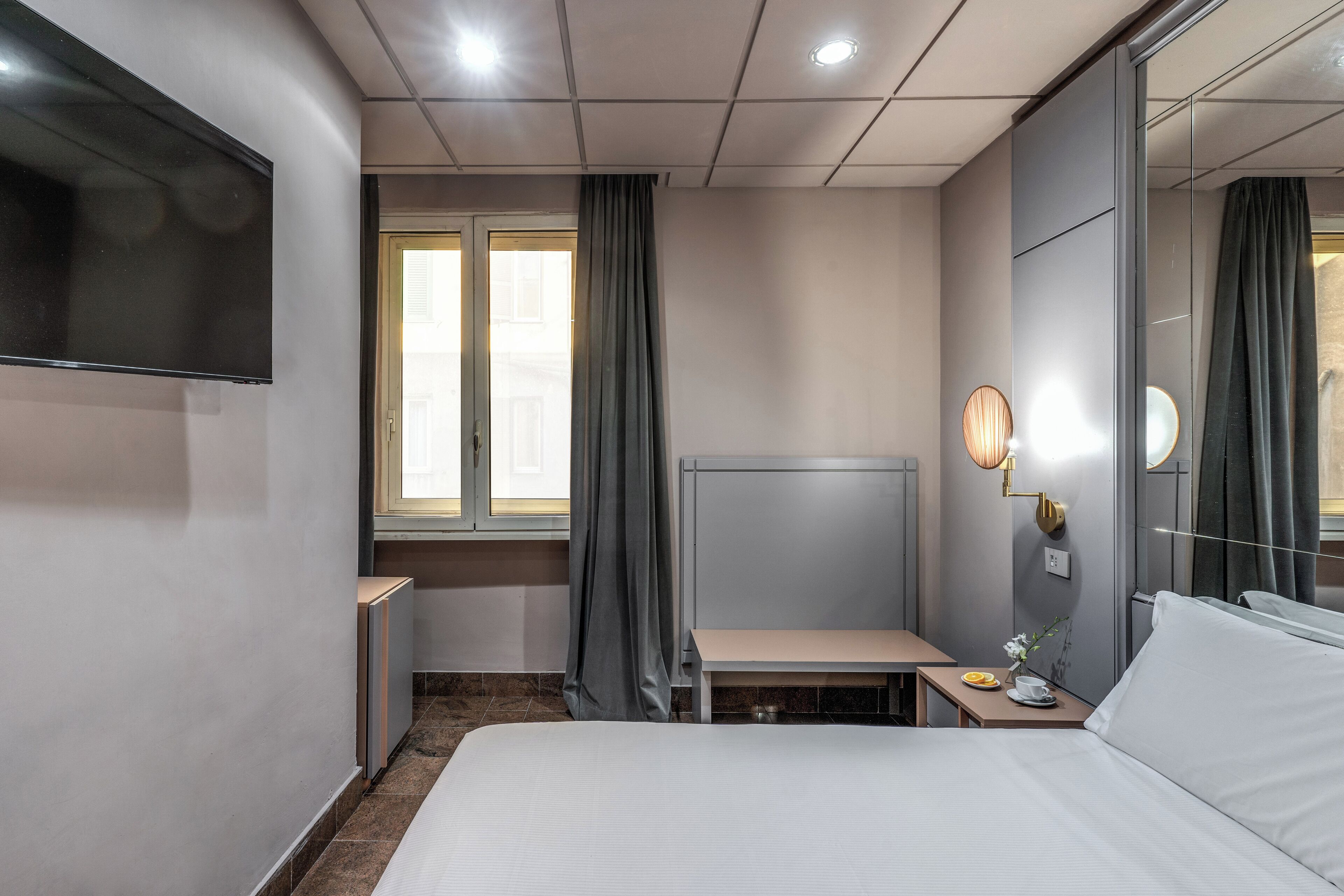 Une chambre d’hôtel comprenant un lit, un bureau, une chaise, une télévision, une table de chevet avec une lampe et une fenêtre avec des rideaux.