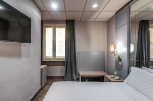 Camera d'albergo con un letto, una scrivania, una sedia, una televisione, un comodino con una lampada e una finestra con tende.