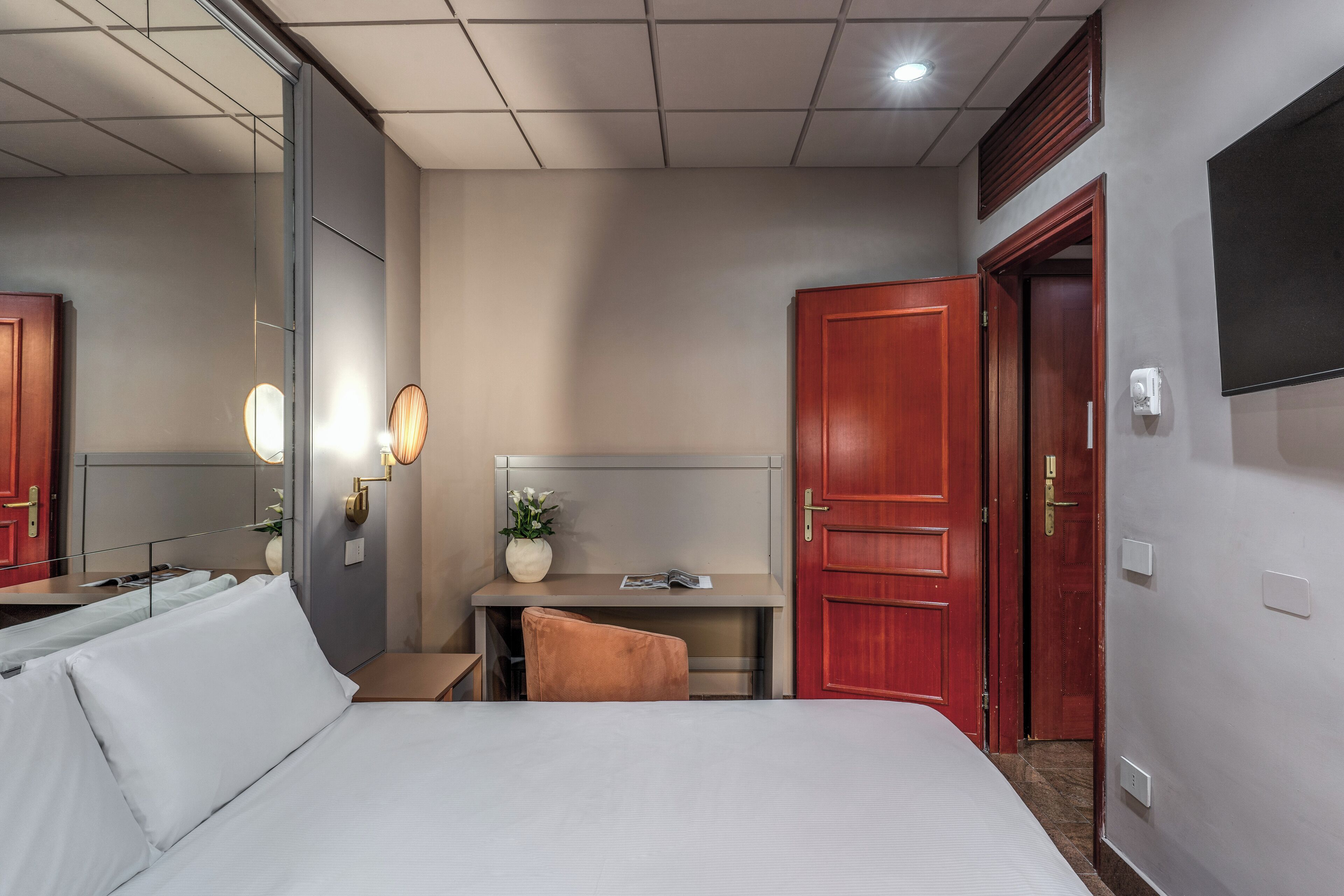 A hotel room with a bed, a desk, a chair, a mirror, and a TV on the wall.