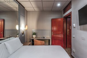 Et hotelværelse med en seng, et skrivebord, en stol, et spejl og et fjernsyn på væggen.