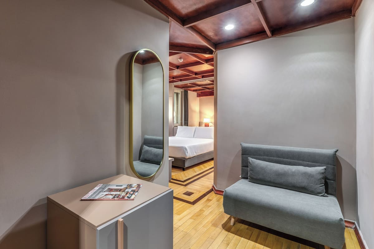 A modern hotel room with a wooden ceiling, a grey sofa, a small table with a magazine, and a large mirror.