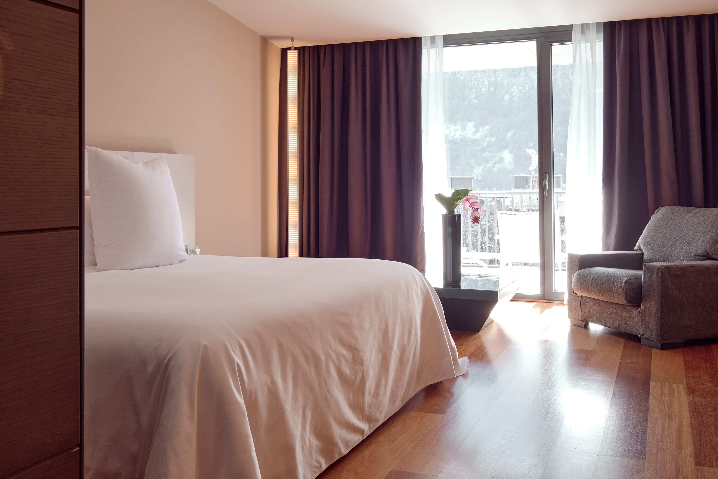 A hotel room with a large bed, a grey armchair, a potted plant, and a view of a balcony with a railing.