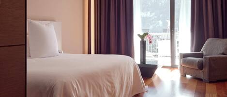 A hotel room with a large bed, a grey armchair, a potted plant, and a view of a balcony with a railing.