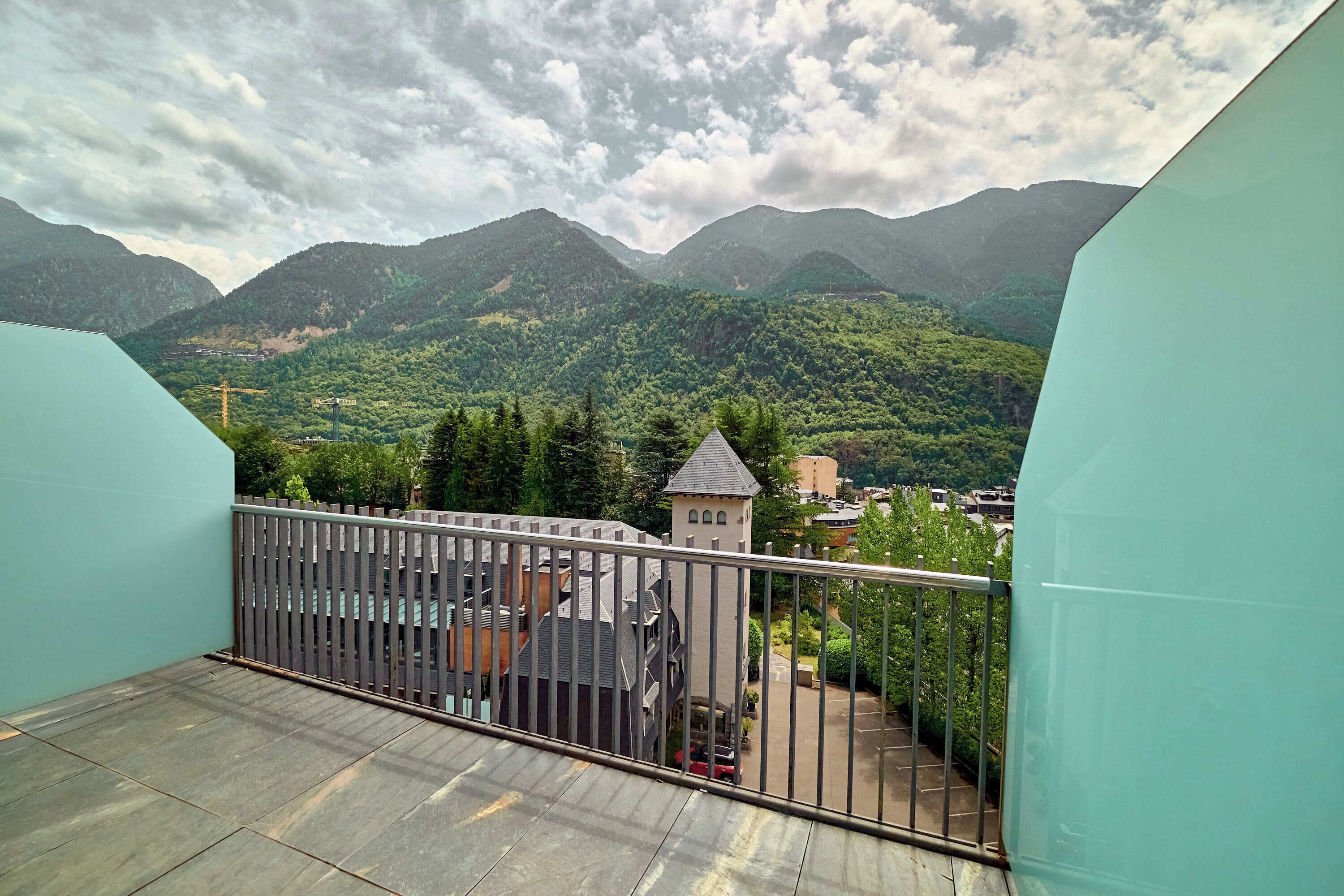 Un balcón con barandilla de metal que da a un paisaje montañoso con una pequeña torre y un aparcamiento.