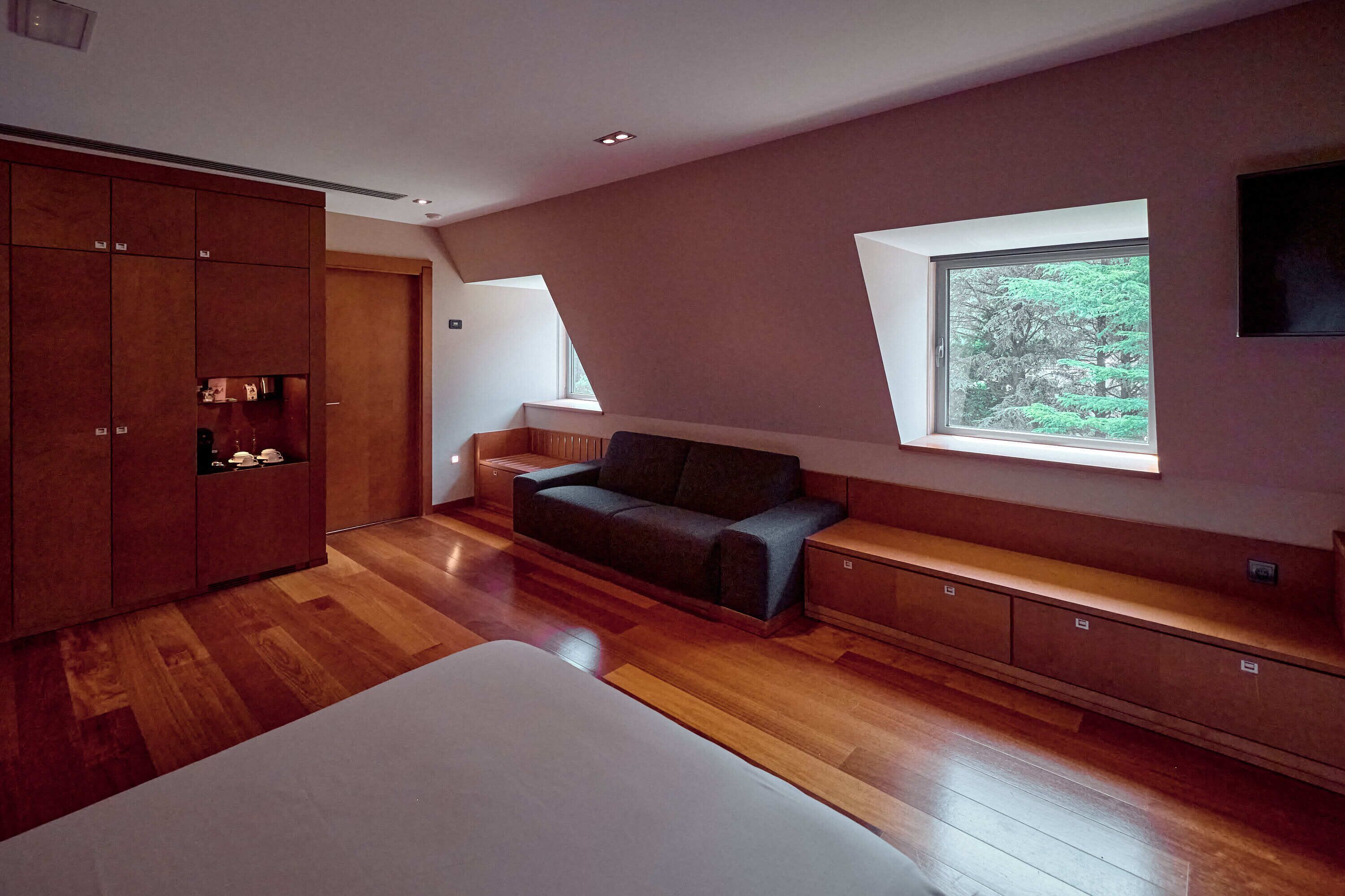 Un dormitorio moderno con suelo de madera, cama, sofá, armario y ventana con vistas a zonas verdes.