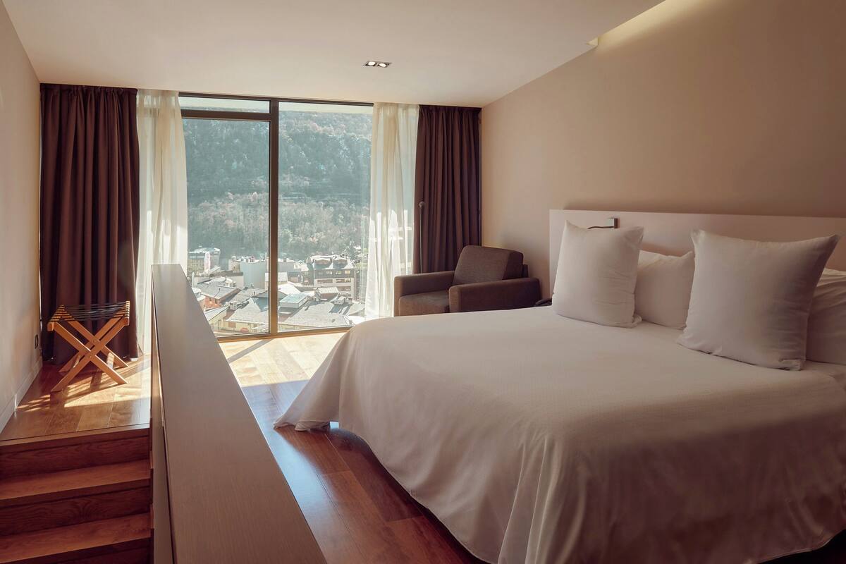 Ein Schlafzimmer mit einem großen Bett, einem Sessel und Blick auf eine gebirgige Landschaft durch ein großes Fenster.