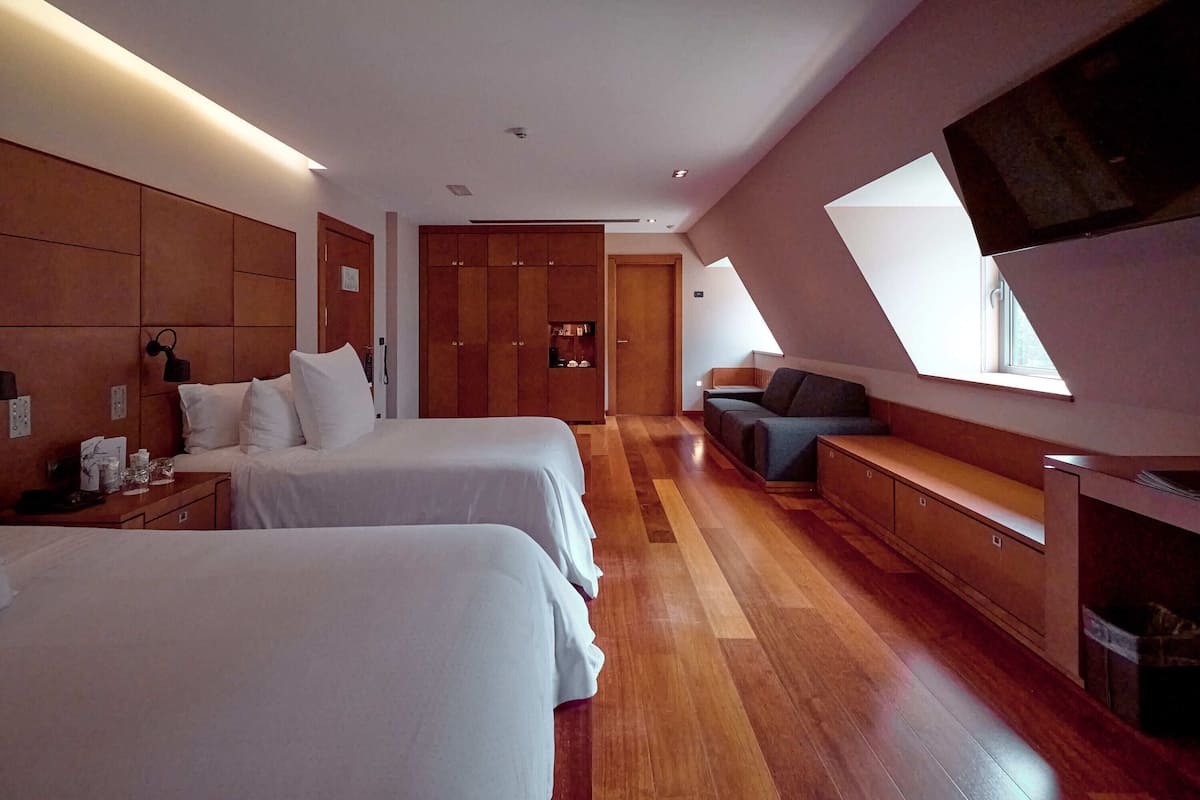 Ein modernes Hotelzimmer mit zwei Betten, einem Holzschrank, einer Sitzecke und einem großen Fenster, das für natürliches Licht sorgt.